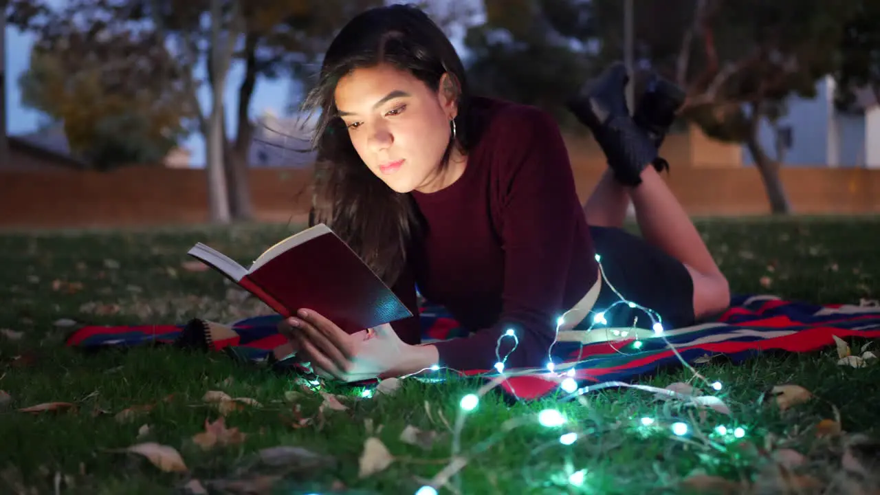 A young hispanic woman student reading a book outdoors at twilight with lights glowing in the night darkness