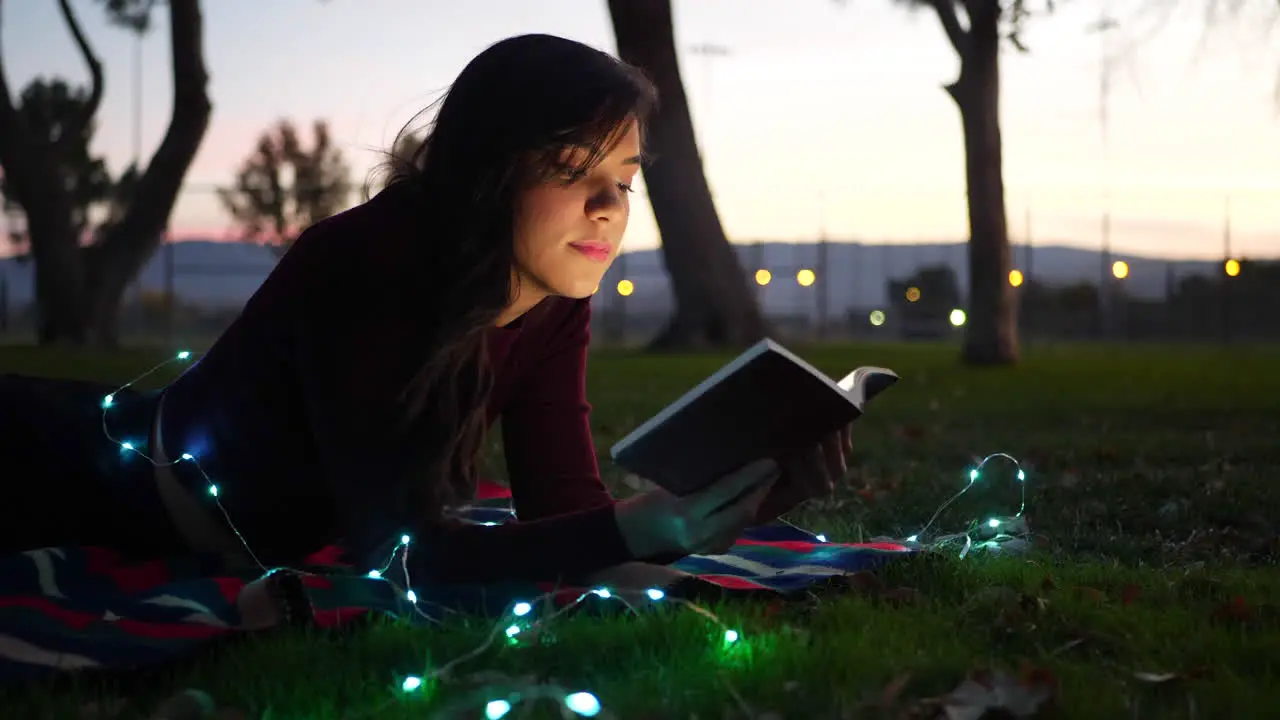 A cute young woman reading a fictional story book outdoors at twilight with lights glowing SLIDE RIGHT