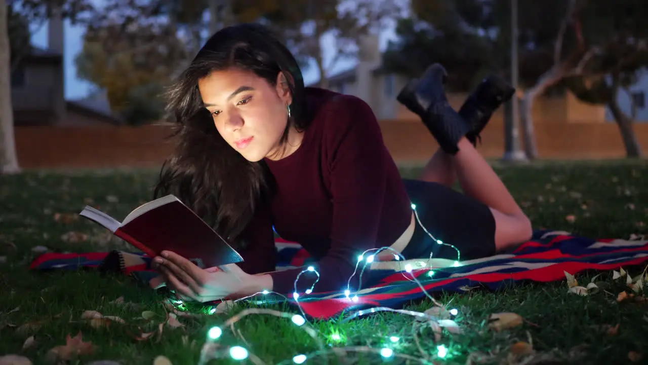 An attractive young woman reading a story book or novel in a park at twilight with lights glowing around her SLIDE LEFT