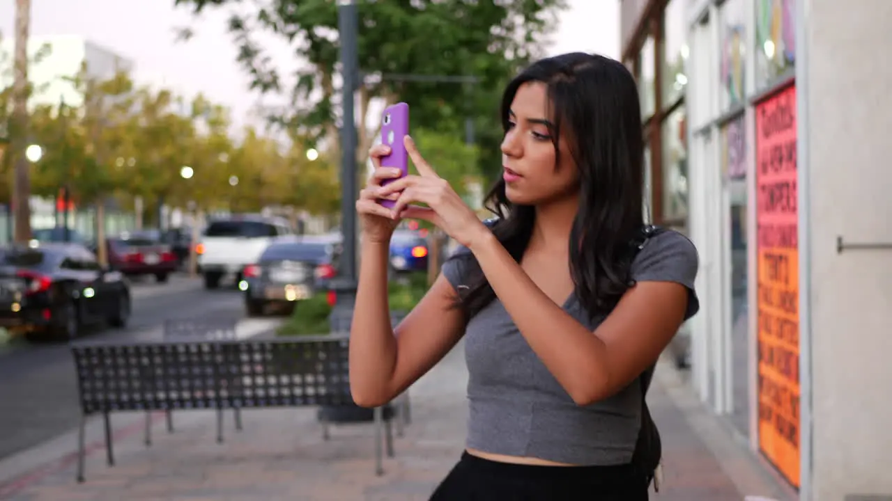 An attractive young hispanic woman walking on the urban city streets doing photography with her smart phone SLOW MOTION