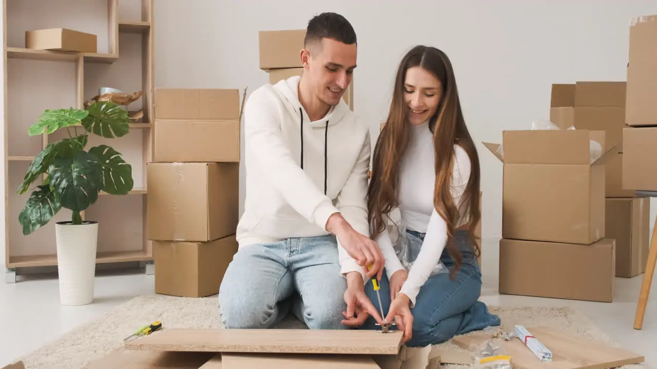 Front View Of A Young Couple In A New House Sitting On The Carpet Assembling A Furniture 1