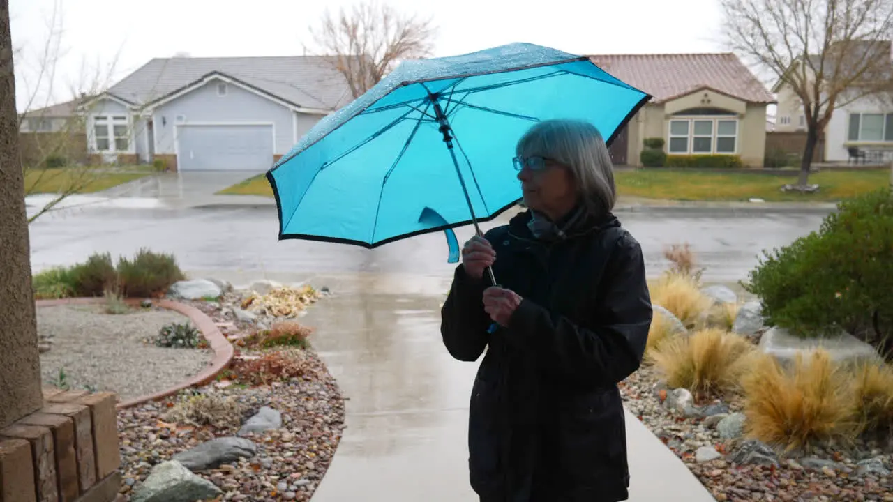 A beautiful old woman walking in the rain and putting away her blue umbrella with raindrops falling in slow motion during a winter storm