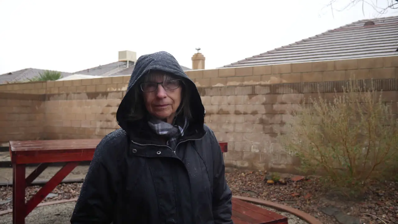 An elderly woman walking in a raincoat and hood during a winter rain storm as drops fall under a cloudy sky in slow motion