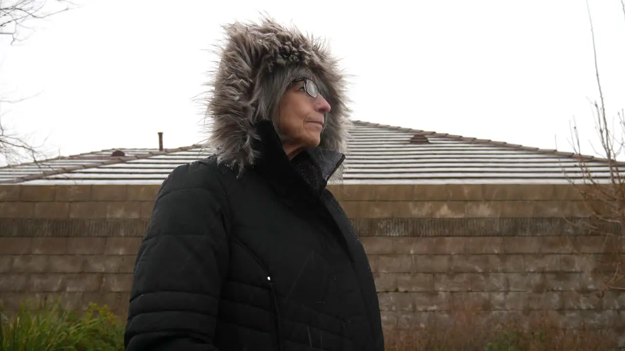 An elderly woman wearing a thick warm winter jacket and hood standing in a freezing cold winter snow storm SLOW MOTION