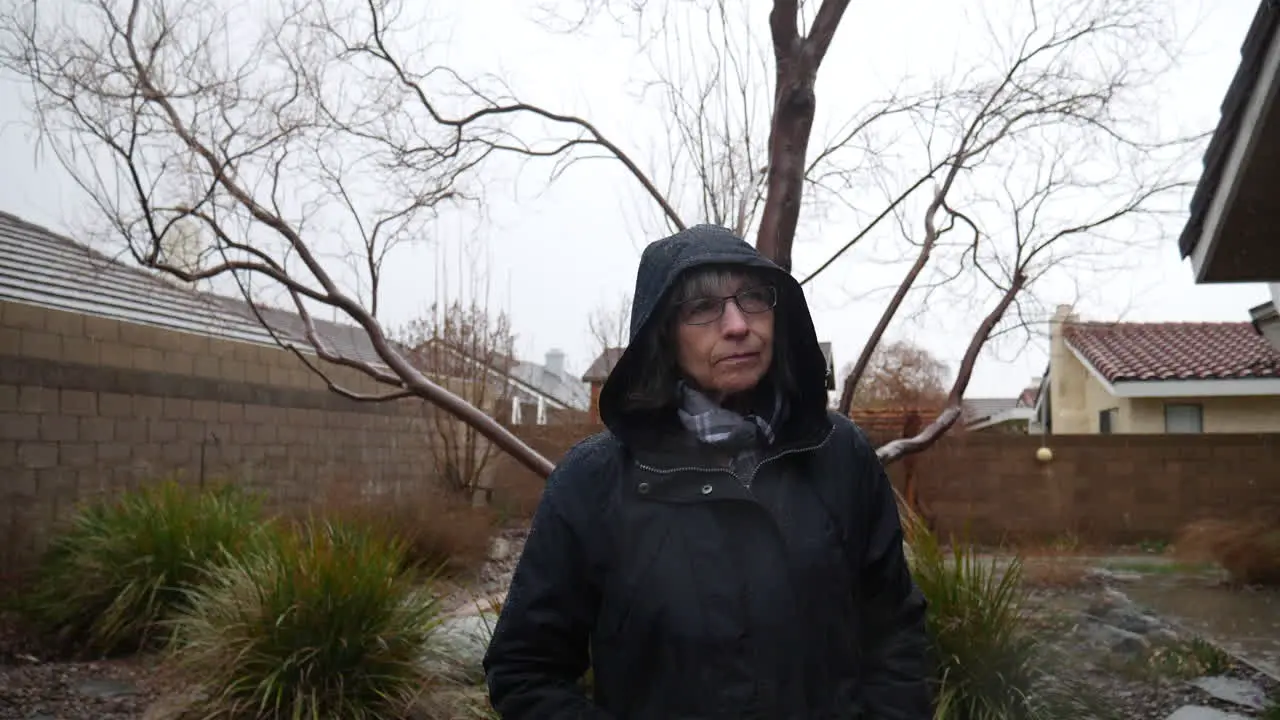 A beautiful old woman wearing a black raincoat and hood during a winter rain storm as drops fall from the cloudy sky in slow motion