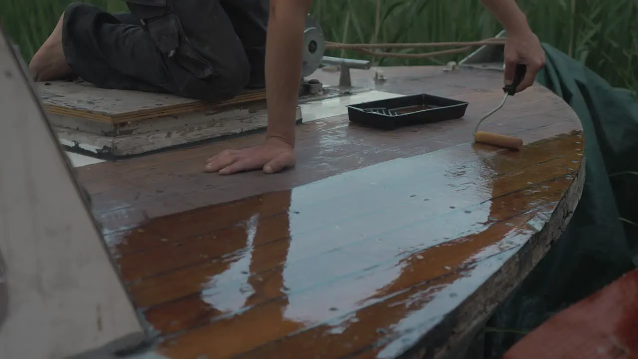 Young man sealing bow of wooden boat deck rolling on varnish