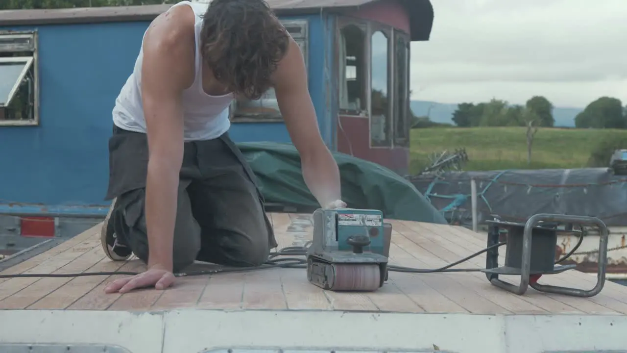 Young carpenter wearing white vest belt sanding cabin roof planking of old wooden boat