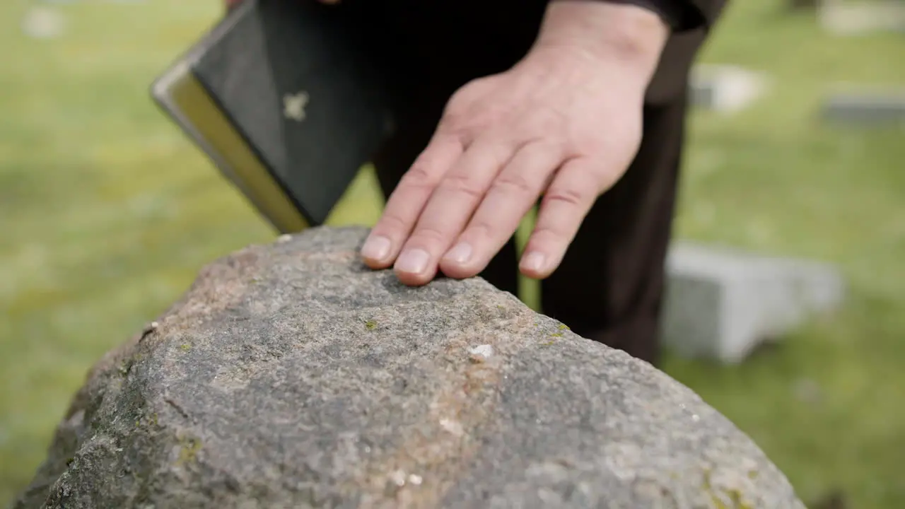 Close Up View Of Man Hand Touching A Tombstone While Holding A Bible In A Graveyard 2