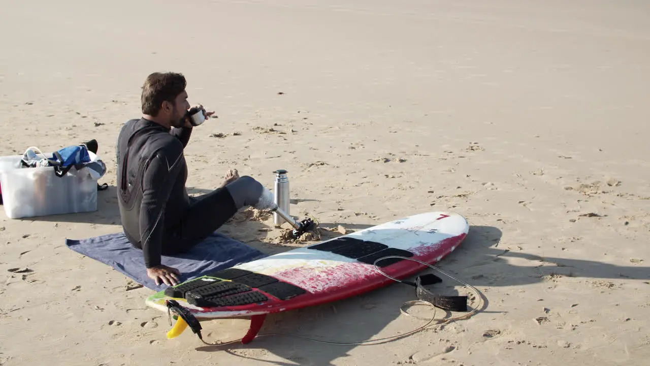 Static Shot Of A Male Surfer With Artificial Leg Drinking Tea On Beach