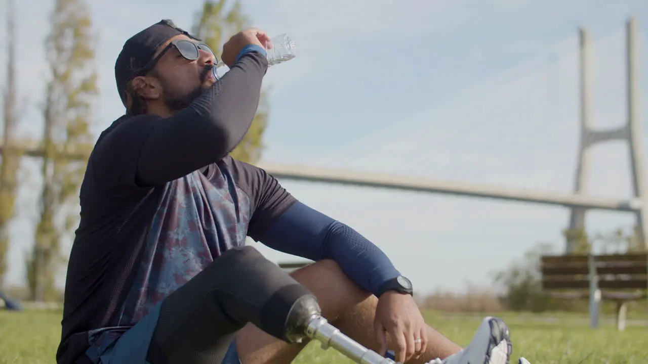 Medium Shot Of A Tired Sportsman With Artificial Leg Drinking Water On The Grass In The Park And Relaxing After Intense Cardio Workout In The Morning