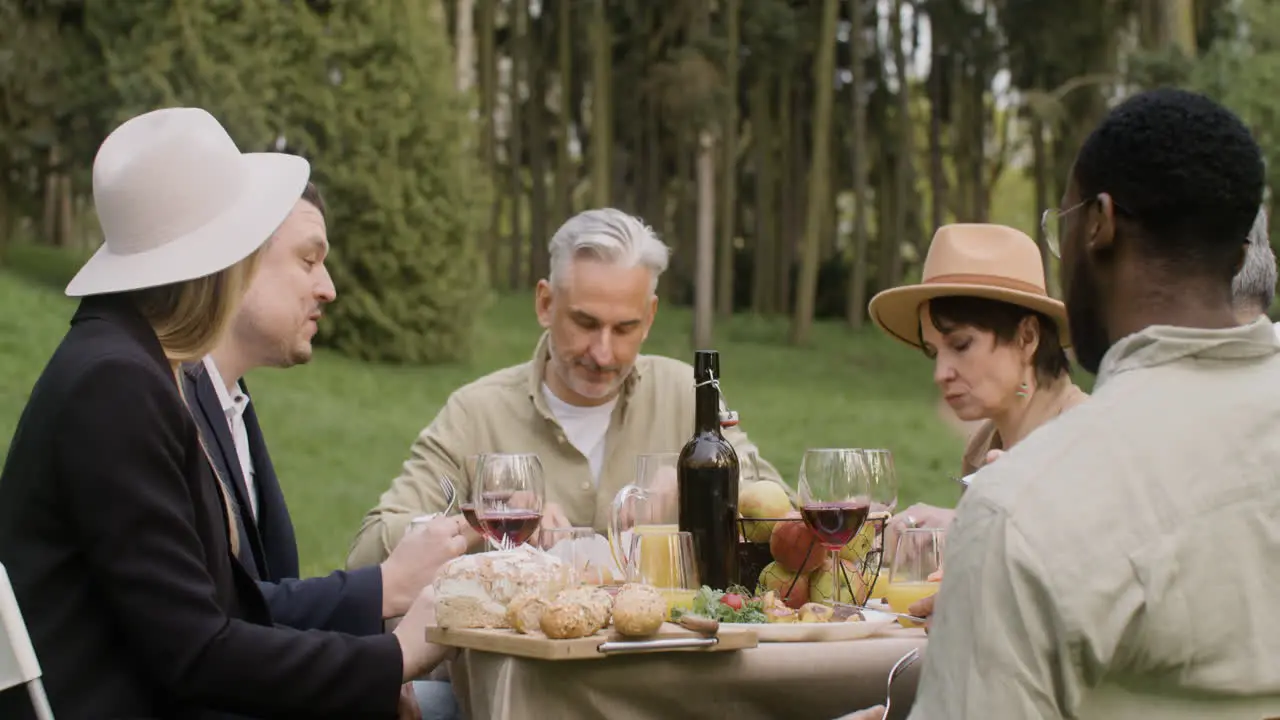 Group Of Middle Aged Friends Eating And Talking To Each Other Sitting At Table During An Outdoor Party In The Park