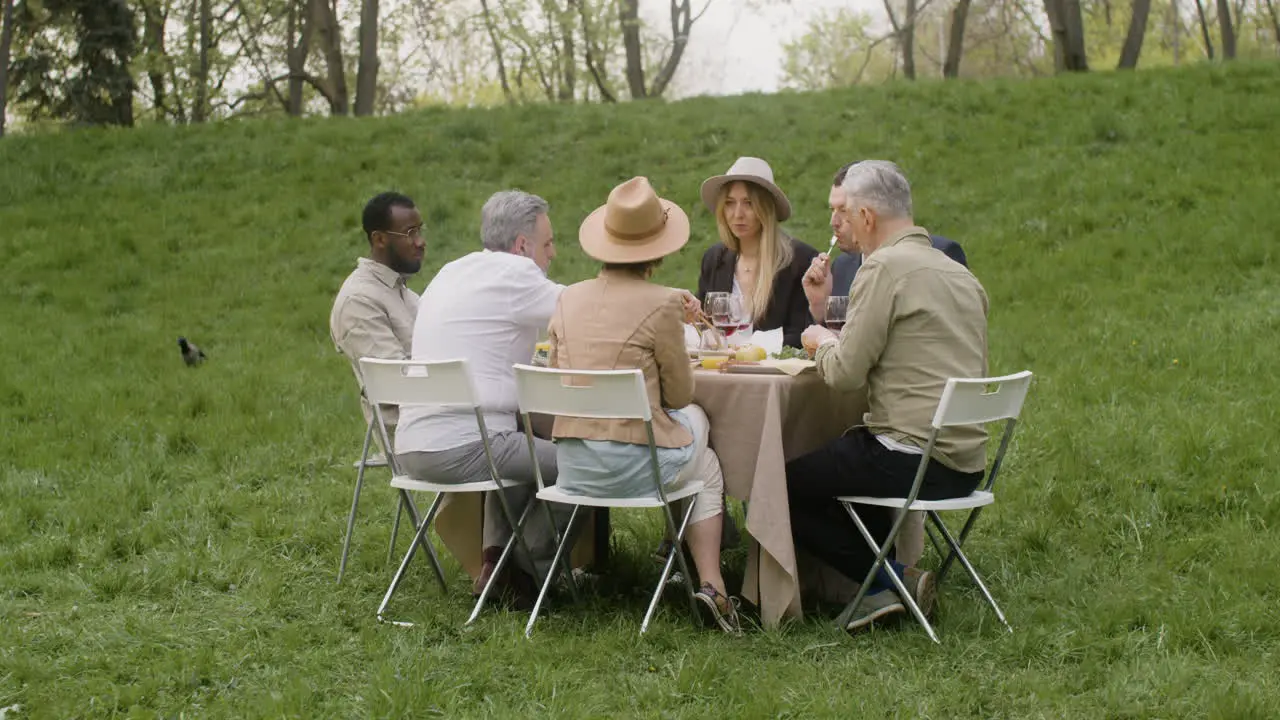 Distant View Of A Group Of Middle Aged Friends Eating And Talking To Each Other Sitting At Table During An Outdoor Party In The Park