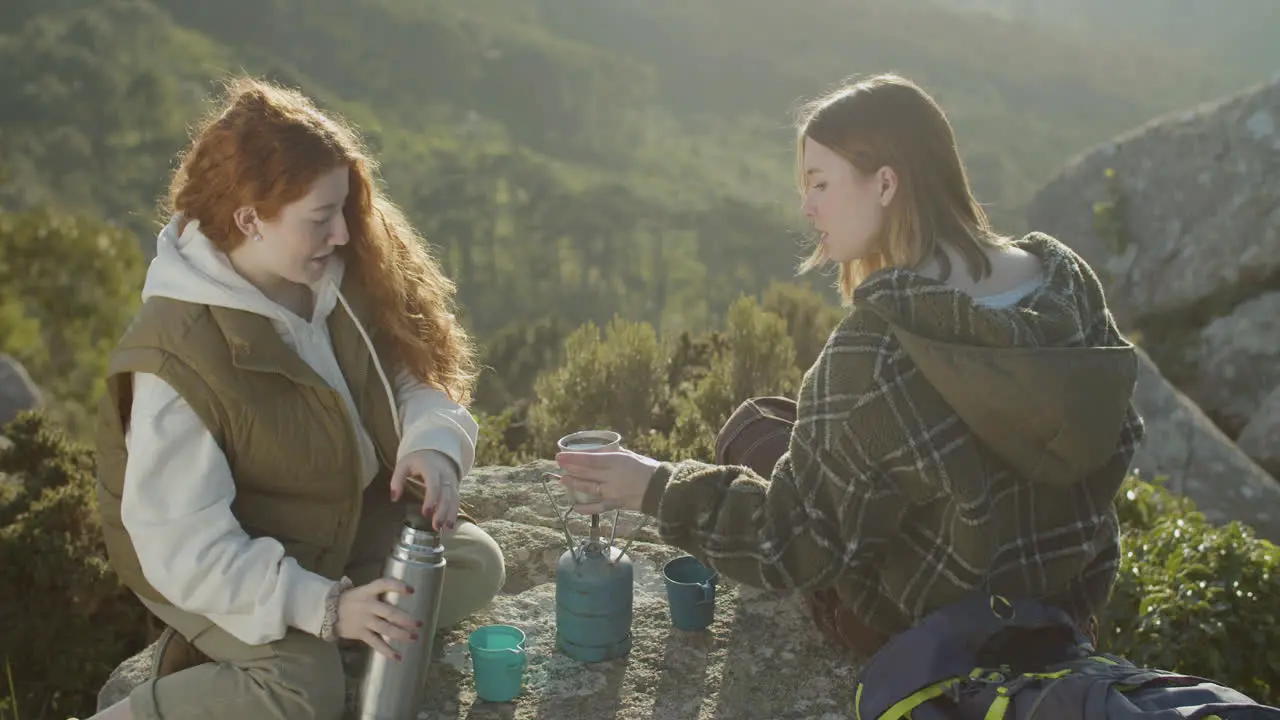 Two Young Female Hikers Drinking Hot Tea From Thermos While Sitting At Mountain Cliff On A Sunny Autumn Day