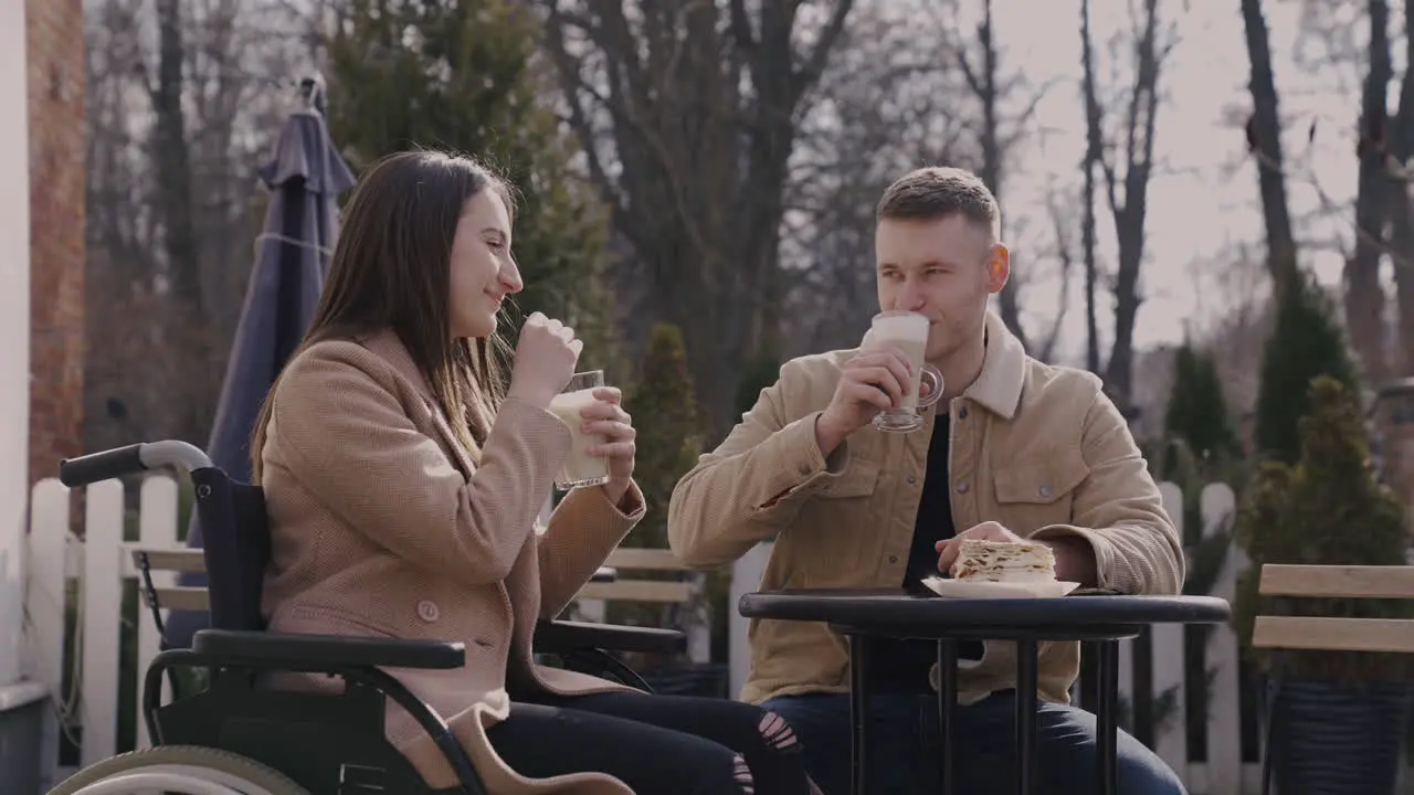 Disabled Girl In Wheelchair And Her Male Friend Drinking Milkshake And Talking Together While Sitting In A Bar Terrace