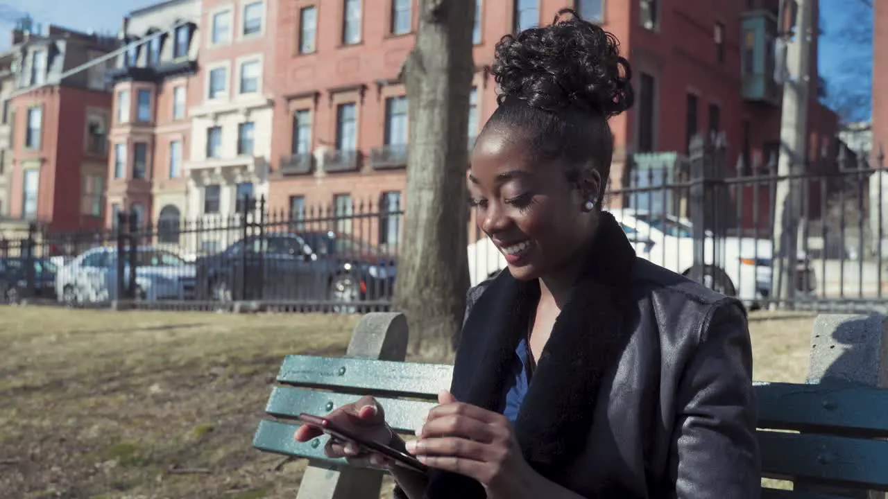 Attractive Young African American Woman Using Smartphone Sitting On Park Bench Receiving Text Message That Makes Her Laugh And Smile Sunny Day In The City