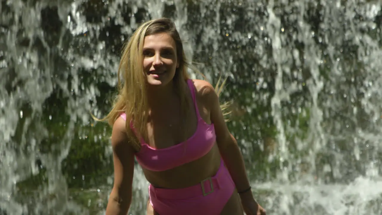 Fun young woman in a bikini in front of waterfall smiles and splashes water at the camera medium shot slow motion