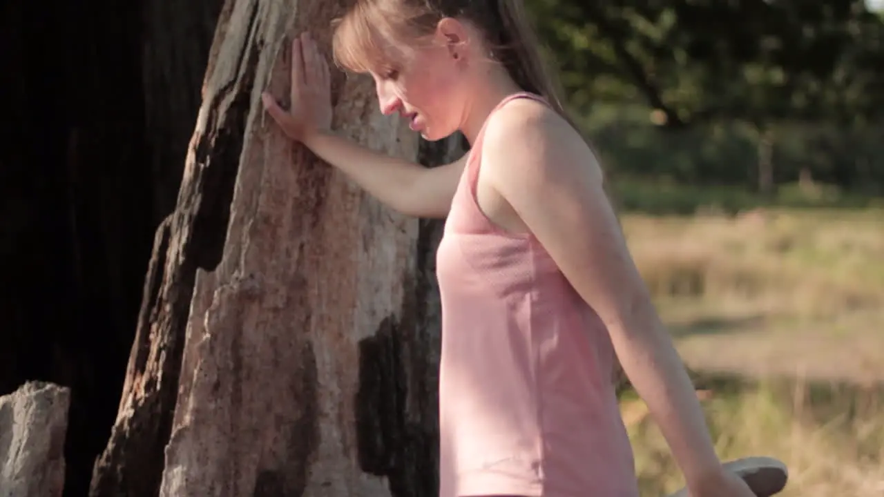 A young woman holds the tree while stretching her leg with her hand before jogging or running in the park on a sunny day