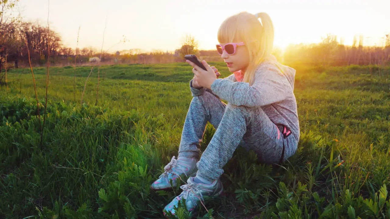 A 6-Year-Old Girl In A Sport Suit Is Sitting On The Grass Using A Mobile Phone