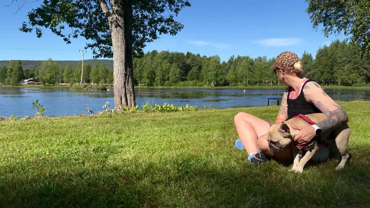 A woman sitting in the green grass petting her French Bulldog in front of a lake on a sunny summer day