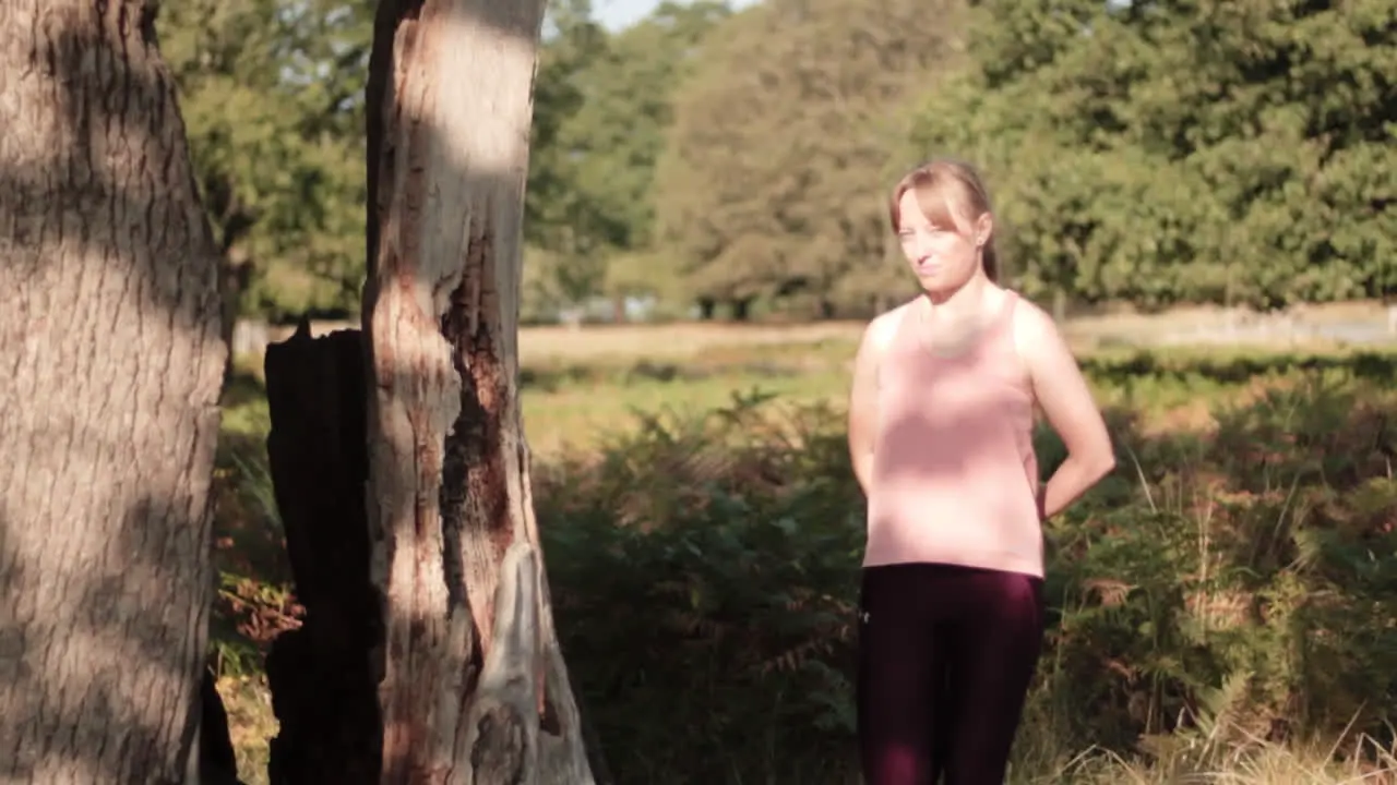 Woman stretching her legs while standing beside the tree for warming up her joints in the park