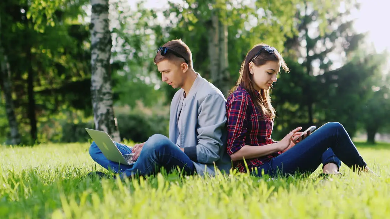 Students Studying In The Park Use A Laptop And A Smartphone They Sit On The Green Grass Hd Video