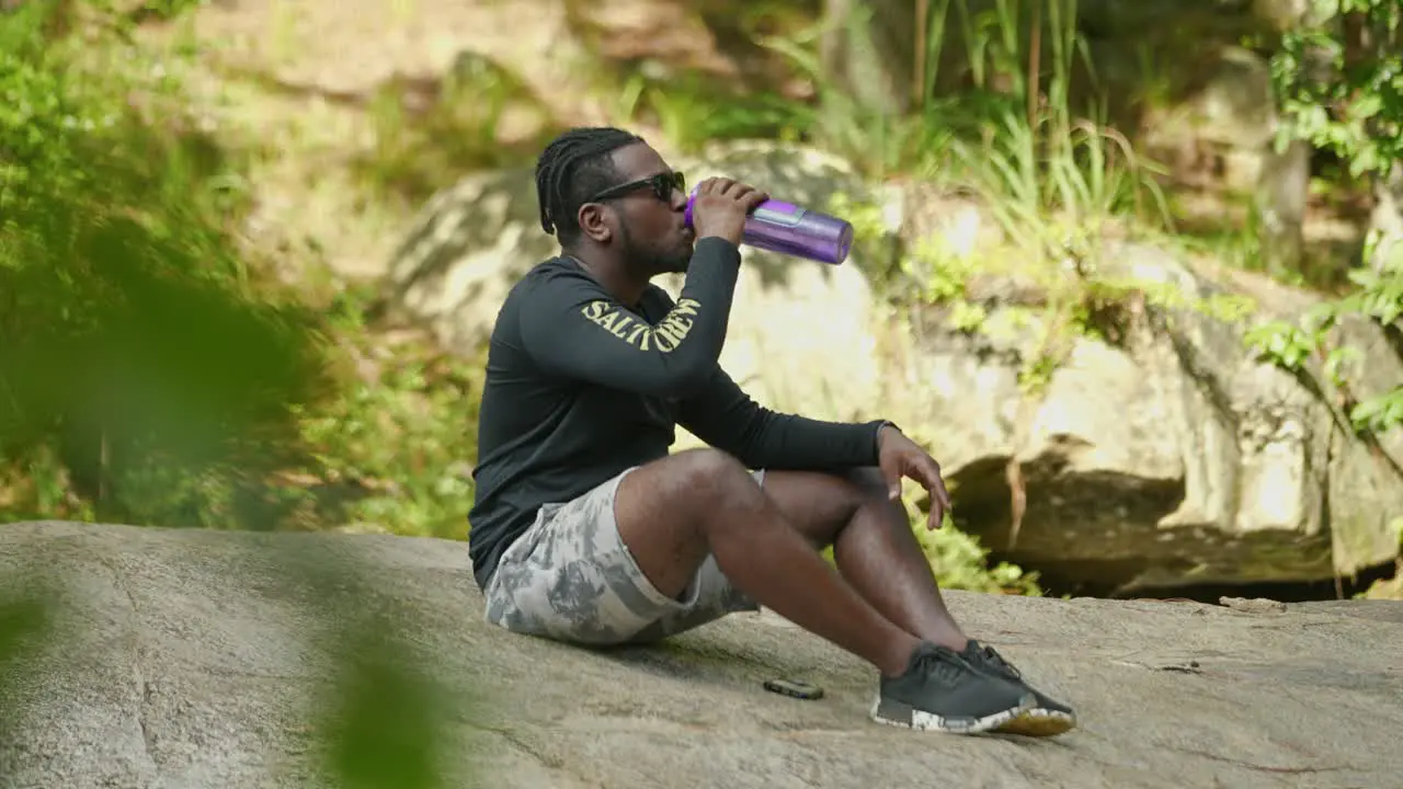 A man takes a sip of water from a bottle on a giant rock in the middle of the woods