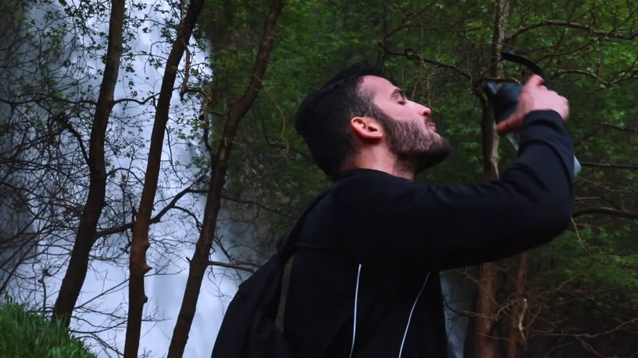 Young thirsty male drinking water in front of a waterfall slow motion medium shot