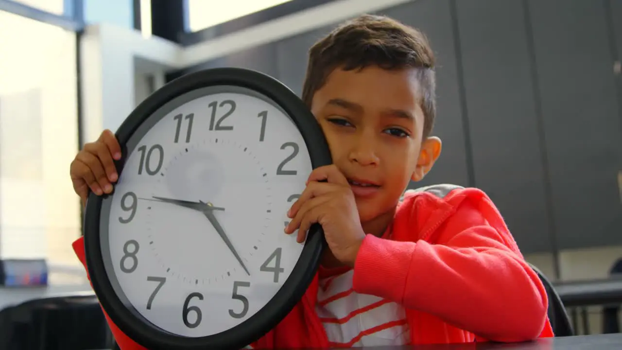 Front view of Asian schoolboy holding wall clock at desk in classroom at school 4k