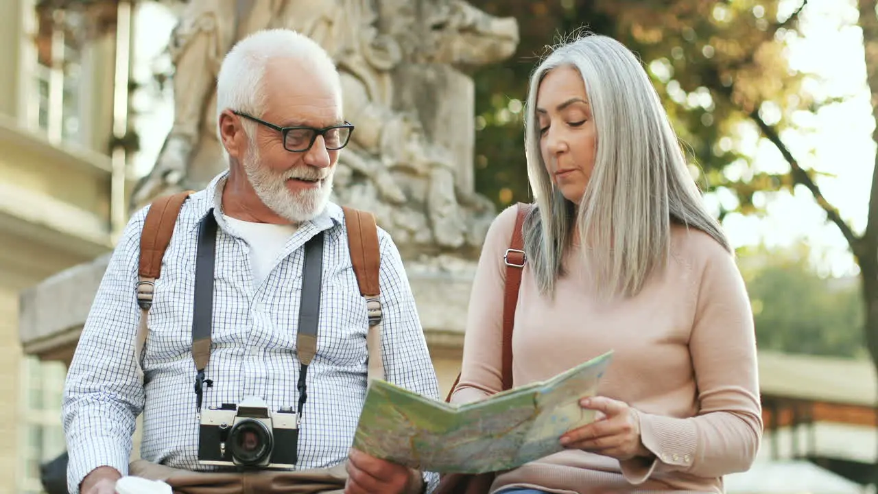 Portrait Of A Couple Of Senior Tourists Sitting On The Monument In The Center Of A City Talking And Looking At The Map