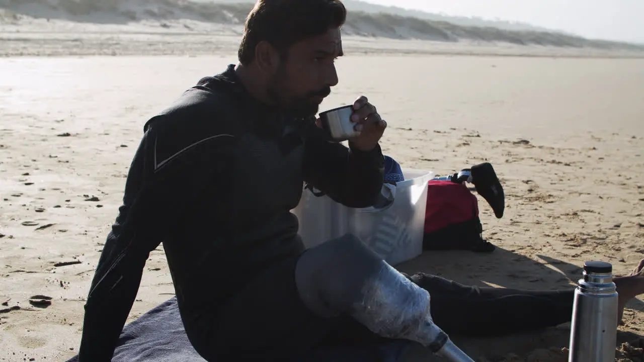 Side View A Male Surfer In Wetsuit With Artificial Leg Drinking Tea On Beach