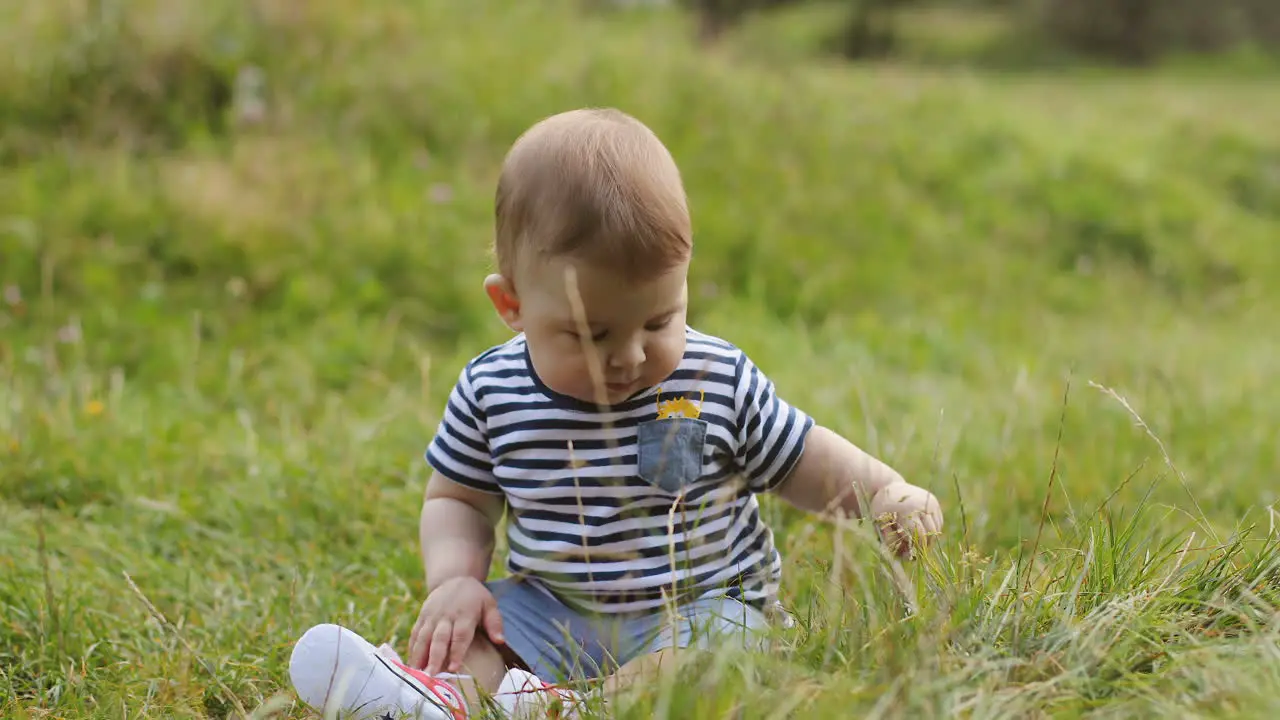 Portrait Of A Lovely Baby Boy Sitting On The Green Grass And Smiling In The Park