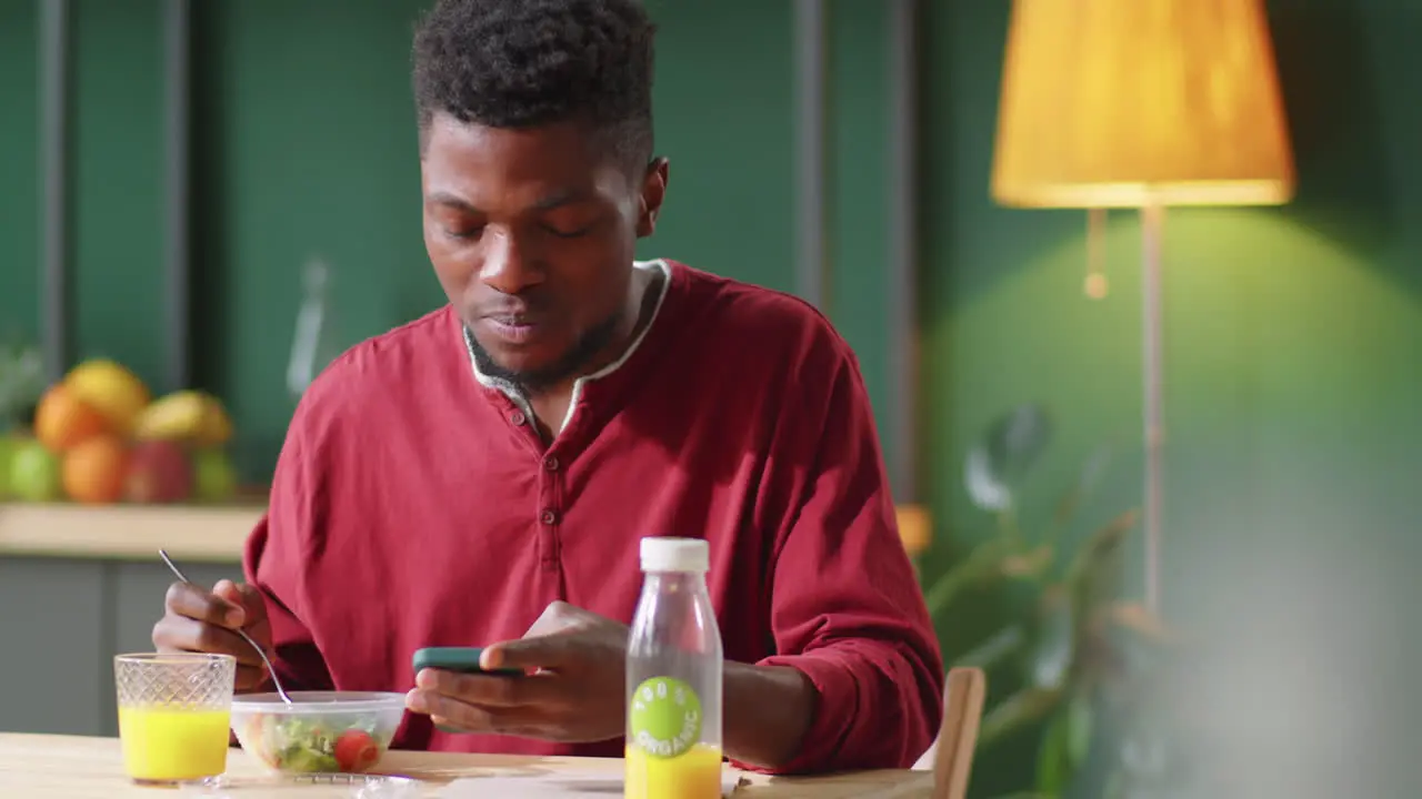 Young Black Man Having Lunch and Using Smartphone at Kitchen Table