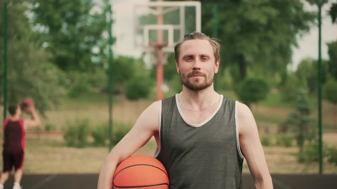 Portrait Of A Handsome Blonde Bearded Basketball Player Holding A Ball While Looking At Camera And Smiling Confidently In An Outdoor Basketball Court