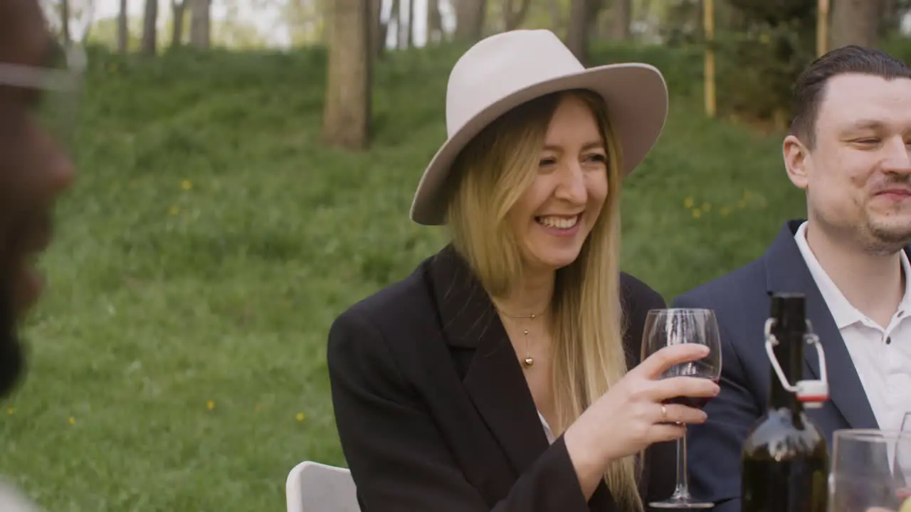 Blond Woman Holding A Red Wine Glass And Laughing While Sitting At Table With Friends During An Outdoor Party In The Park