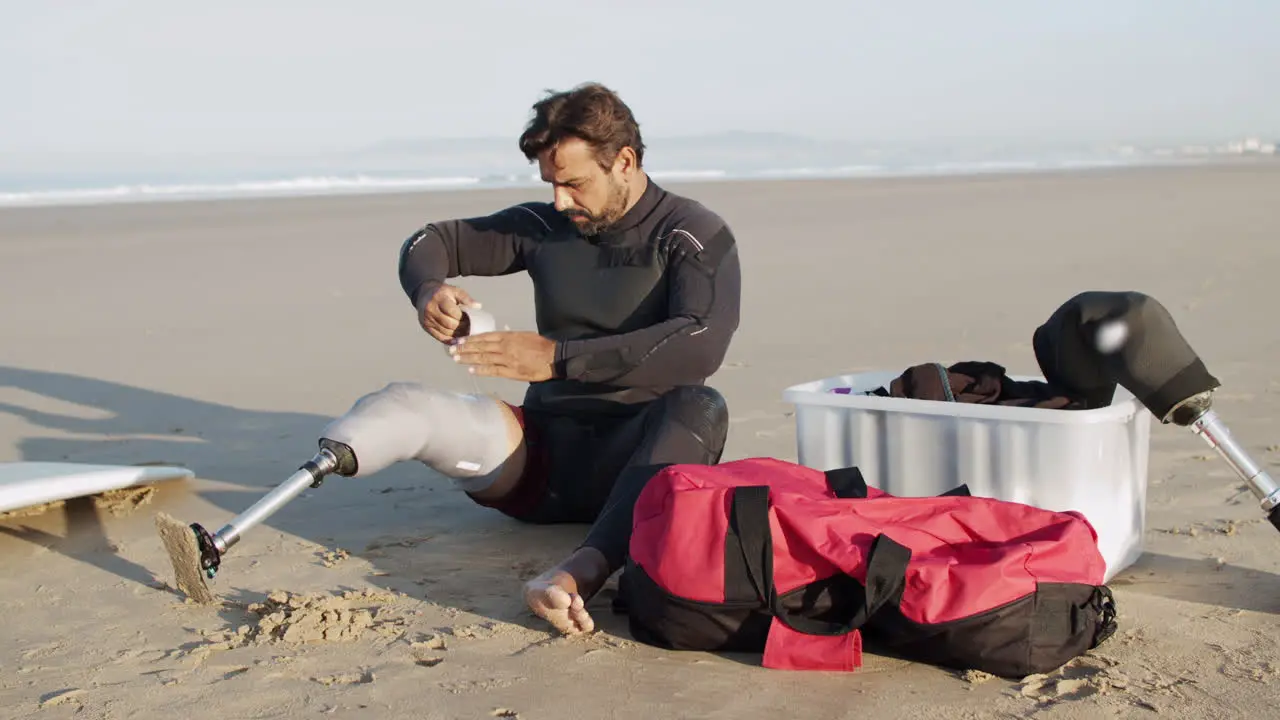 Long Shot Of Male Surfer With Disability Taping Artificial Leg On The Coast