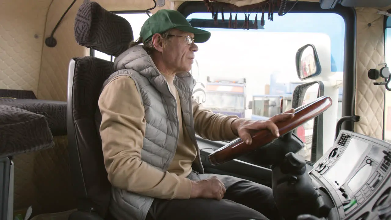 Side View Of Older Worker Wearing Cap And Vest Driving A Truck In A Logistics Park