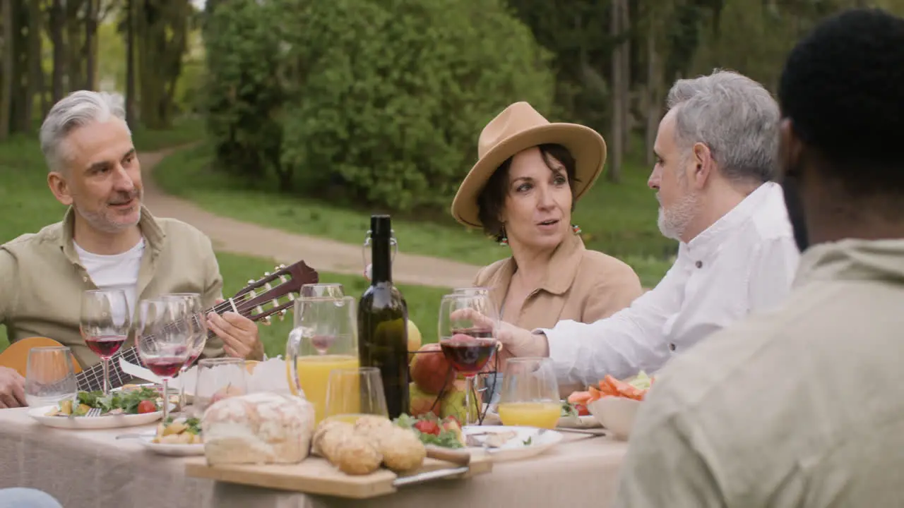 Middle Aged Man Plating A Guitar Sitting At Table And Talking With His Friends During An Outdoor Party In The Park