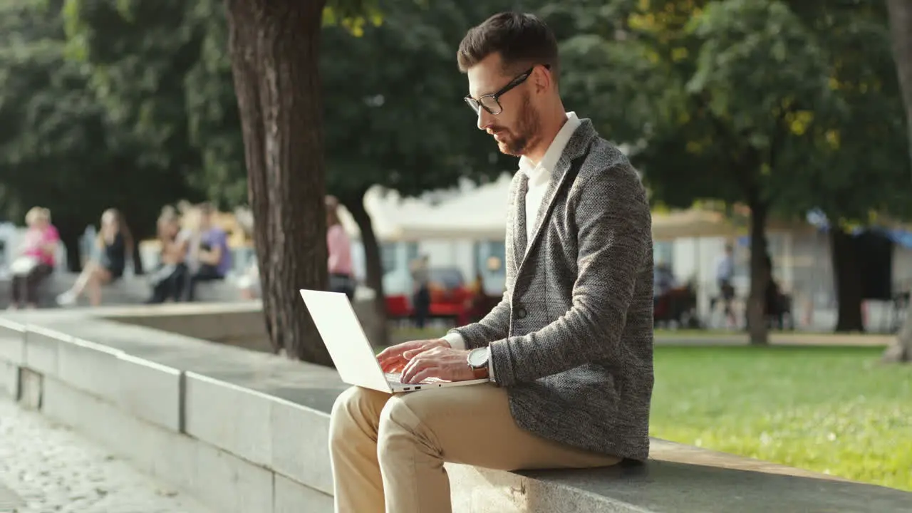 Handsome Businessman Sitting On Wall In The City Park And Working On His Laptop Computer