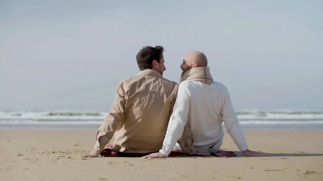 Back View Of A Cheerful Homosexual Couple Leaning Their Heads To Each Other While Sitting On The Beach