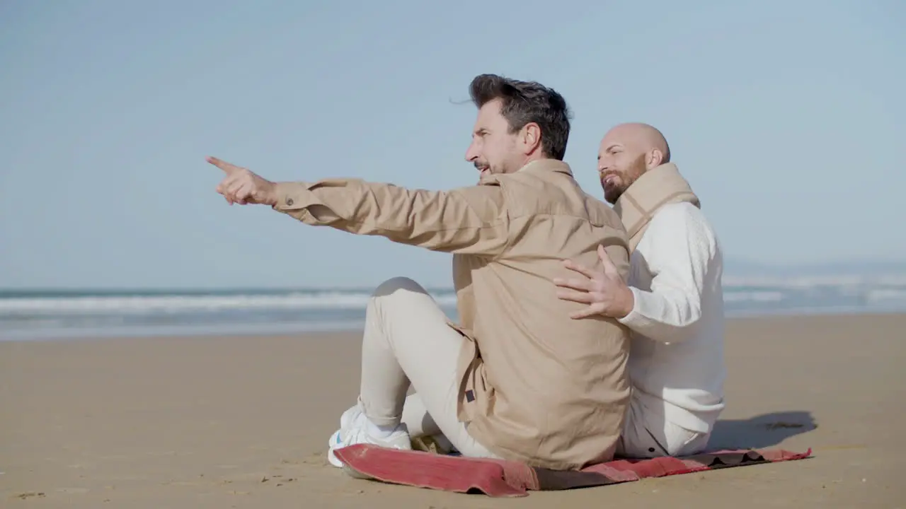 Happy Gay Man Sitting On The Sandy Beach With His Partner And Pointing Finger Into Distance