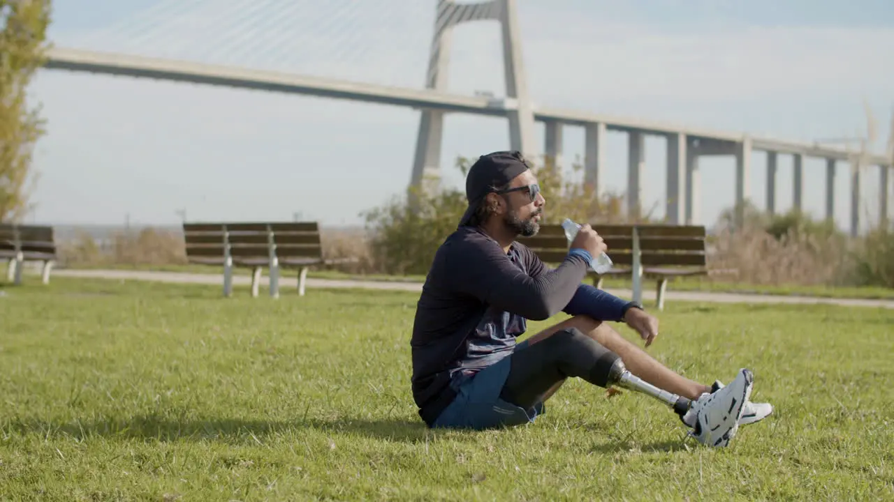 Long Shot Of A Tired Sportsman With Artificial Leg Drinking Water On The Grass In The Park And Relaxing After Intense Cardio Workout In The Morning