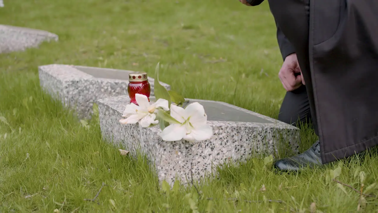 Unrecognizable Man In Black Suit Kneeling And Putting Flowers And A Candle On Tombstone In A Graveyard 1