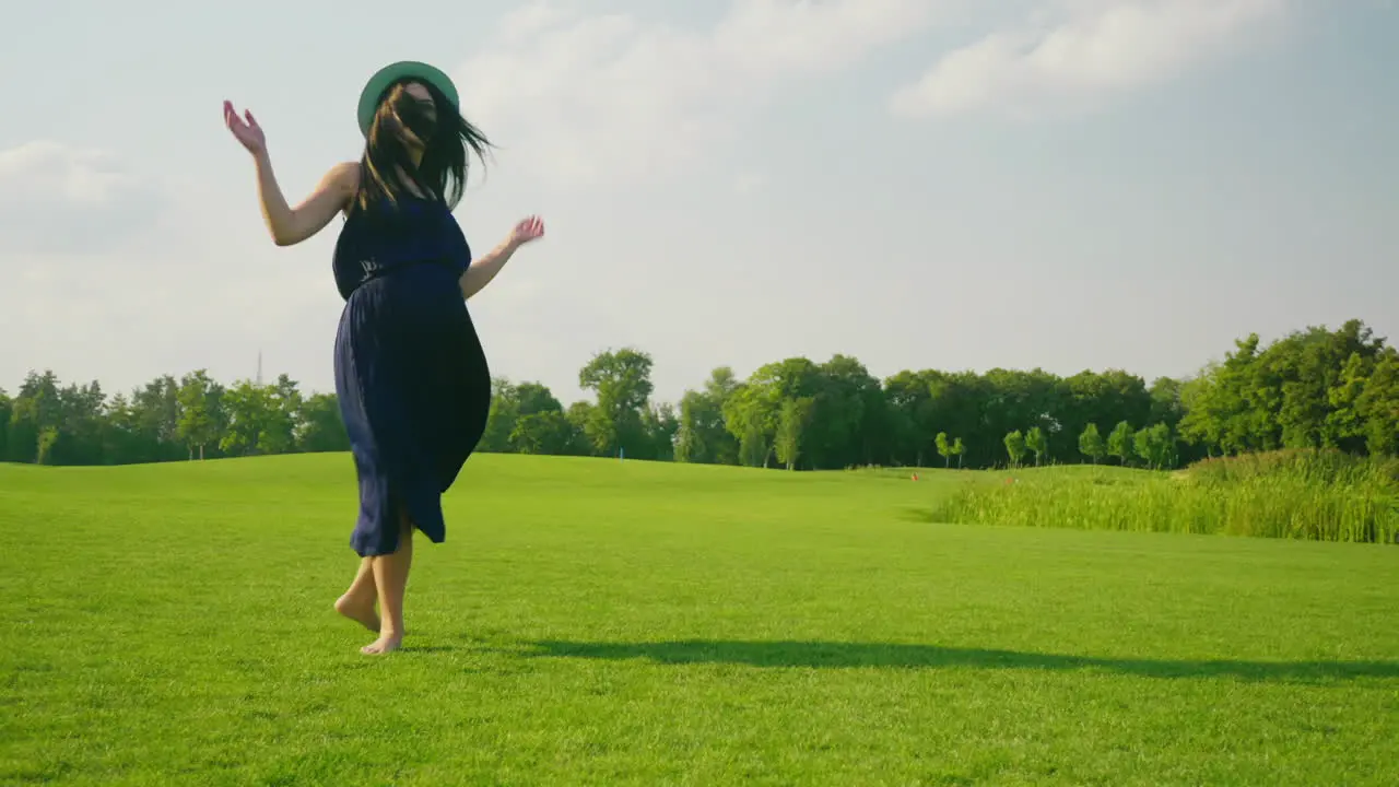 A Stylish Pregnant Woman In A Light Dress Carefree Walks Along A Green Meadow Past The Camera