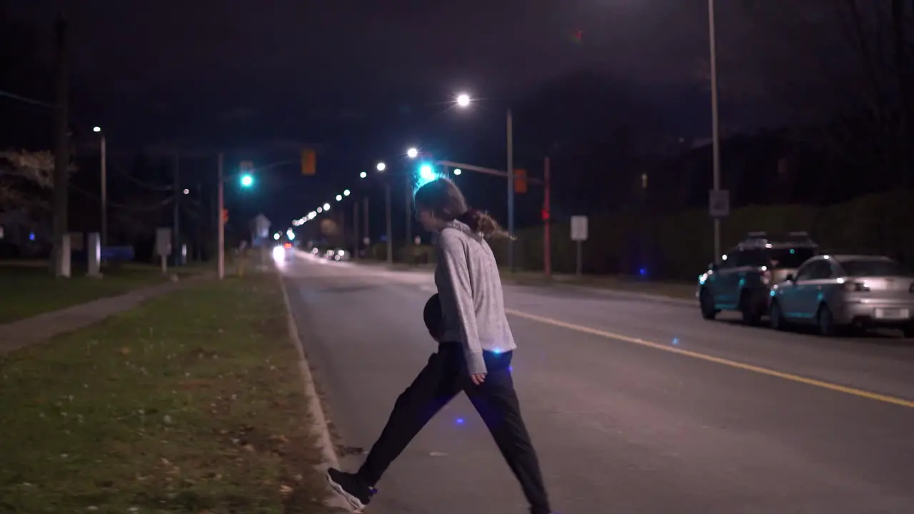 Teenage girl in grey sweater with black hair crosses road with basketball in hand at night