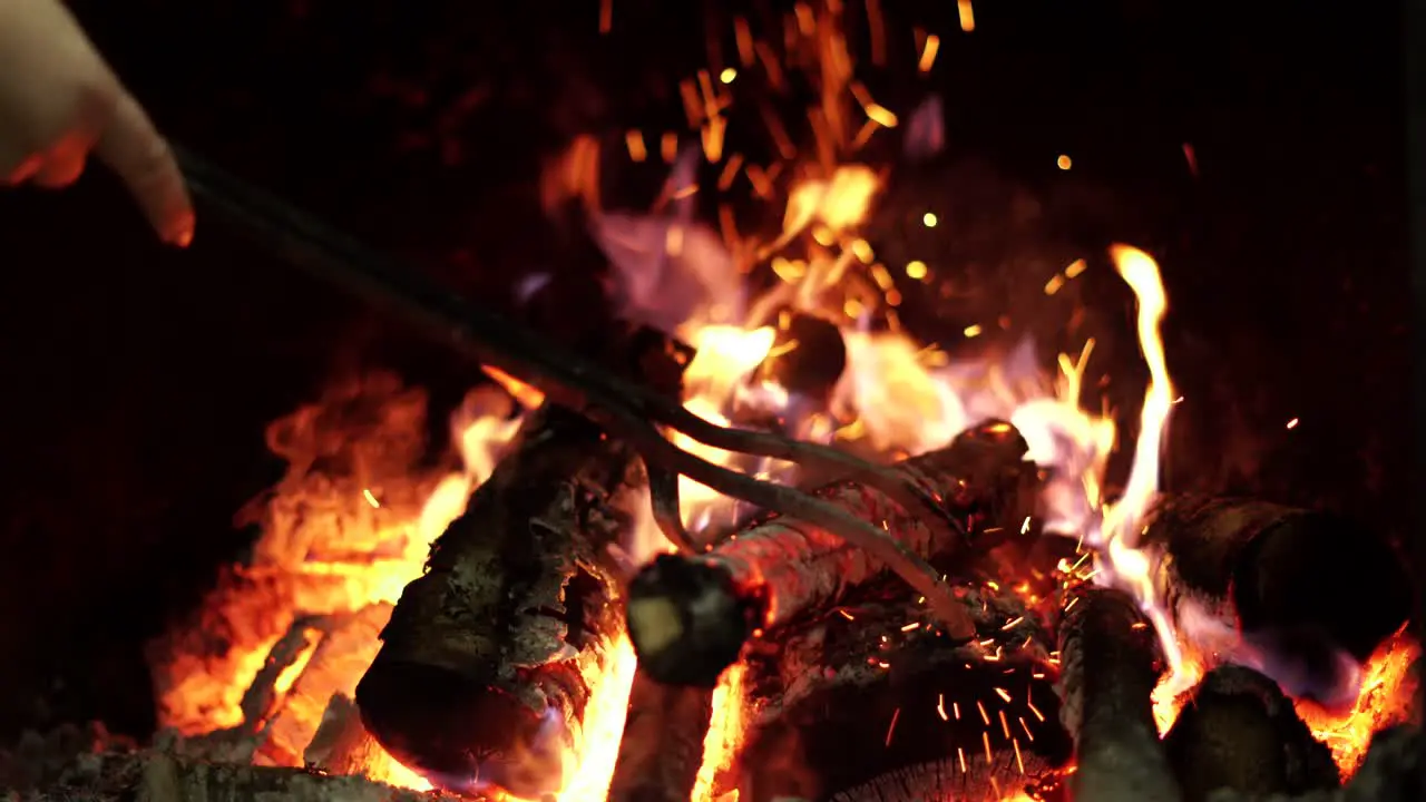 Closeup on fire-iron placing burning logs in open indoor fireplace flames on black background