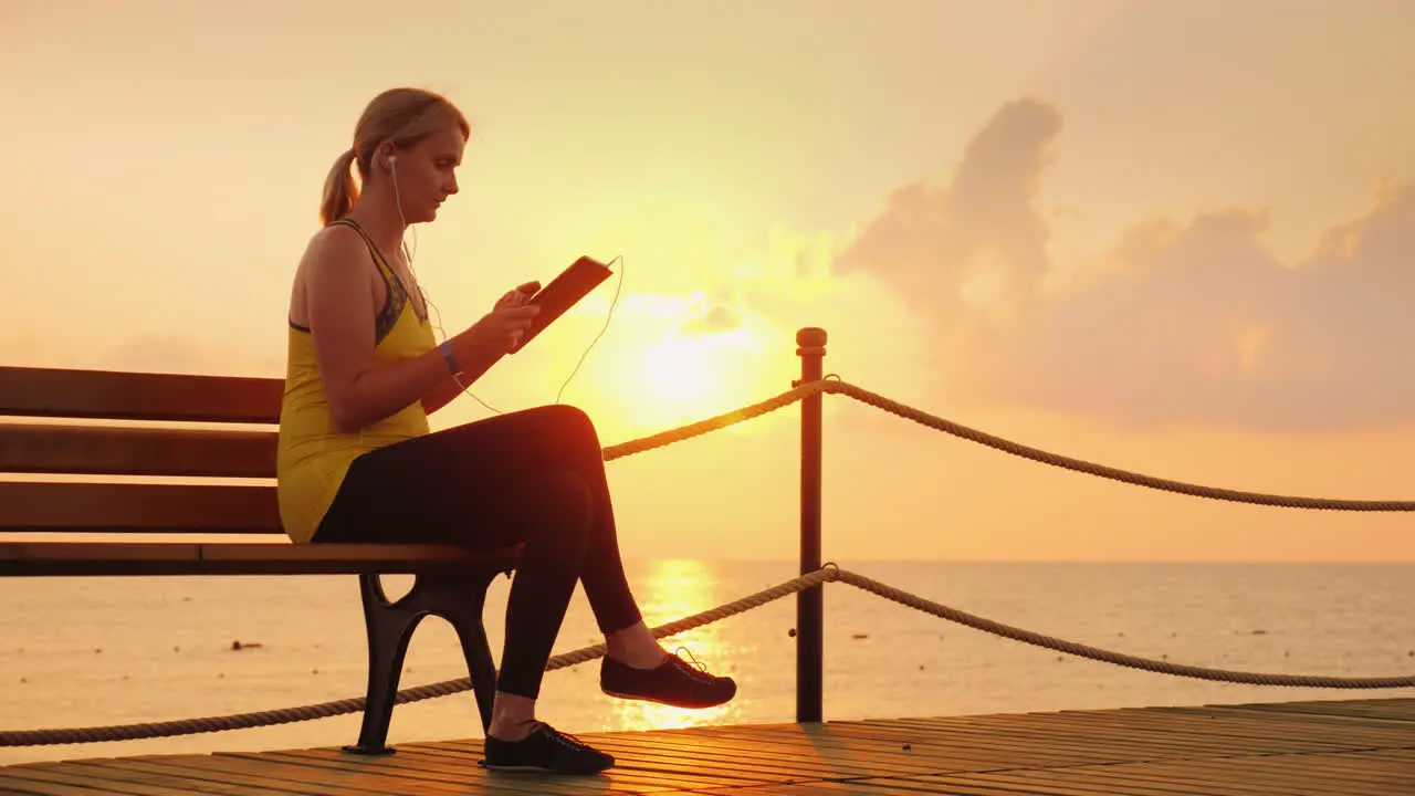 Fitness Woman Enjoys A Tablet Sits On A Bench On A Pier Against The Backdrop Of The Rising Sun 4k Vi