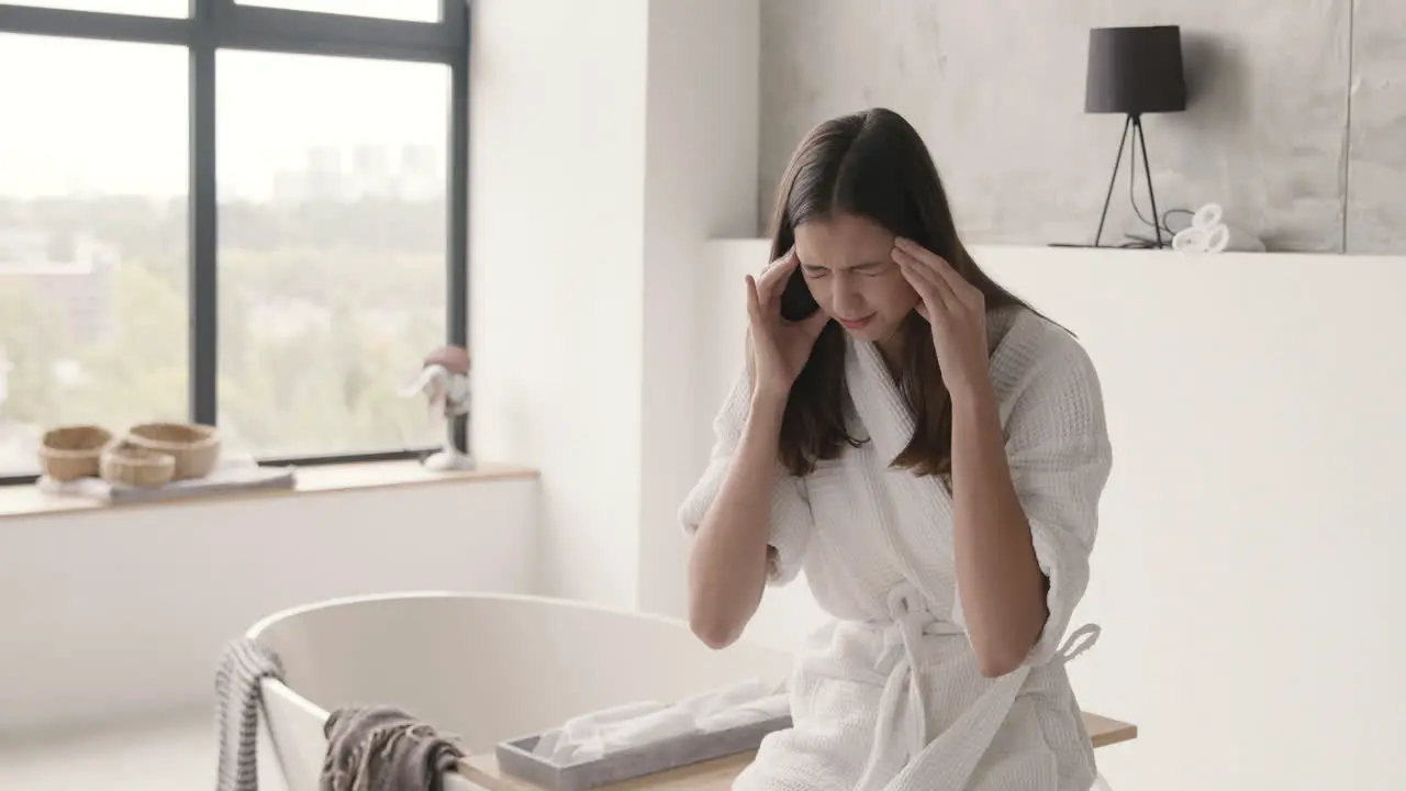 Suffering Woman In Robe Having Headache While Sitting On Bathtub In A Modern Bathroom