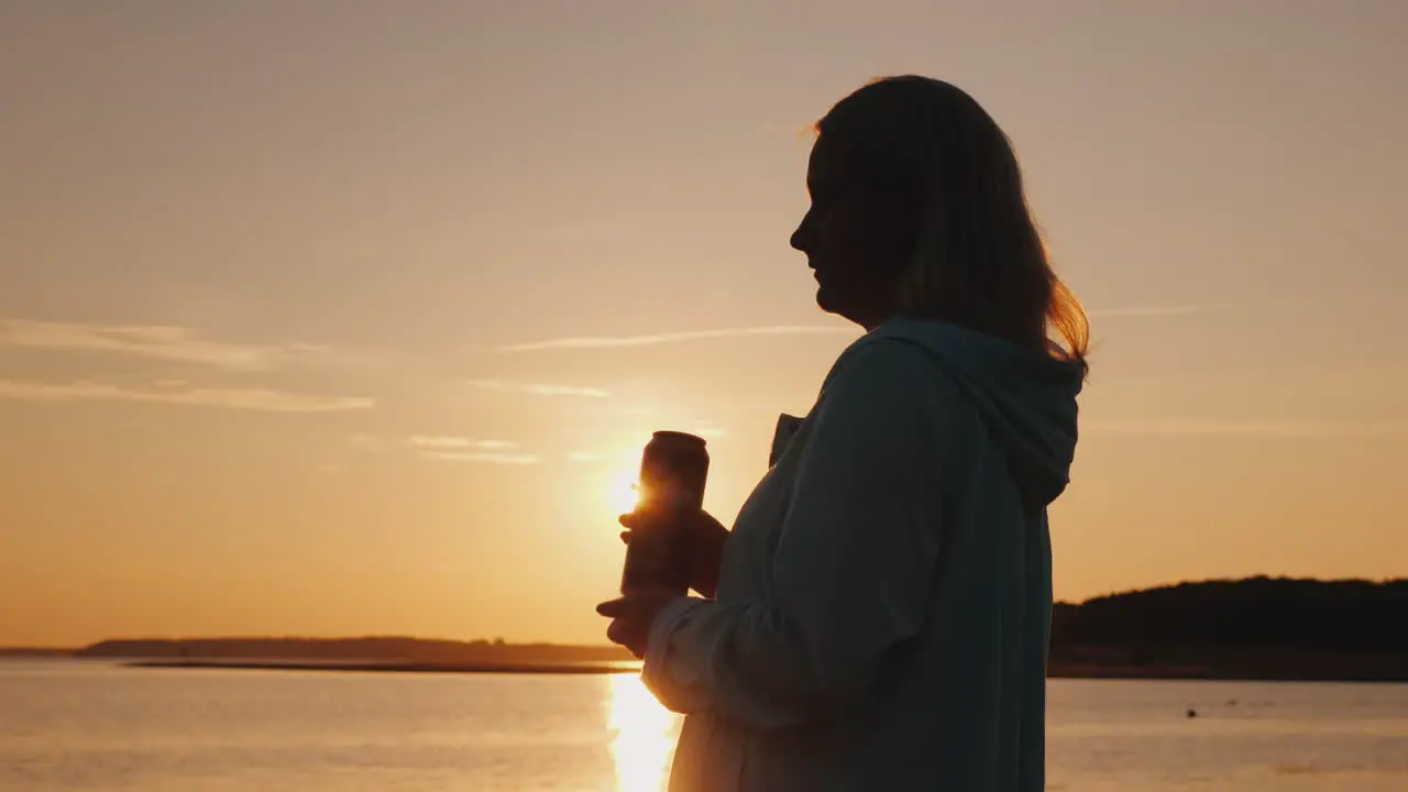 Lonely Woman Drinks Beer From A Can On The Lake Standing Alone By The Fence Looking At The Sunset