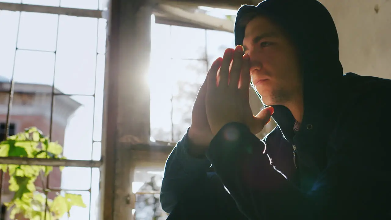 A Young Man In A Hood Sitting On A Window Praying Or Very Thoughtful In The Rays Of The Sun