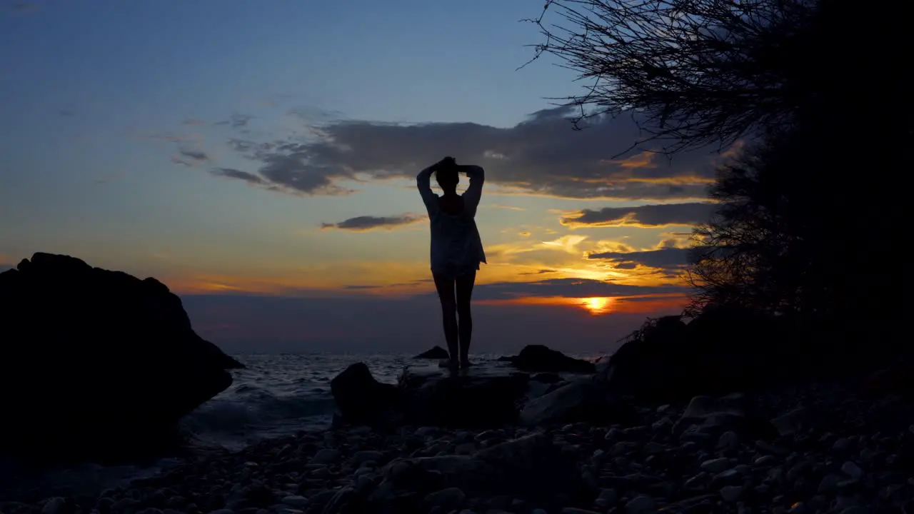 Young woman relaxing on rocky shore watching sea under colorful sky at sunset in Mediterranean